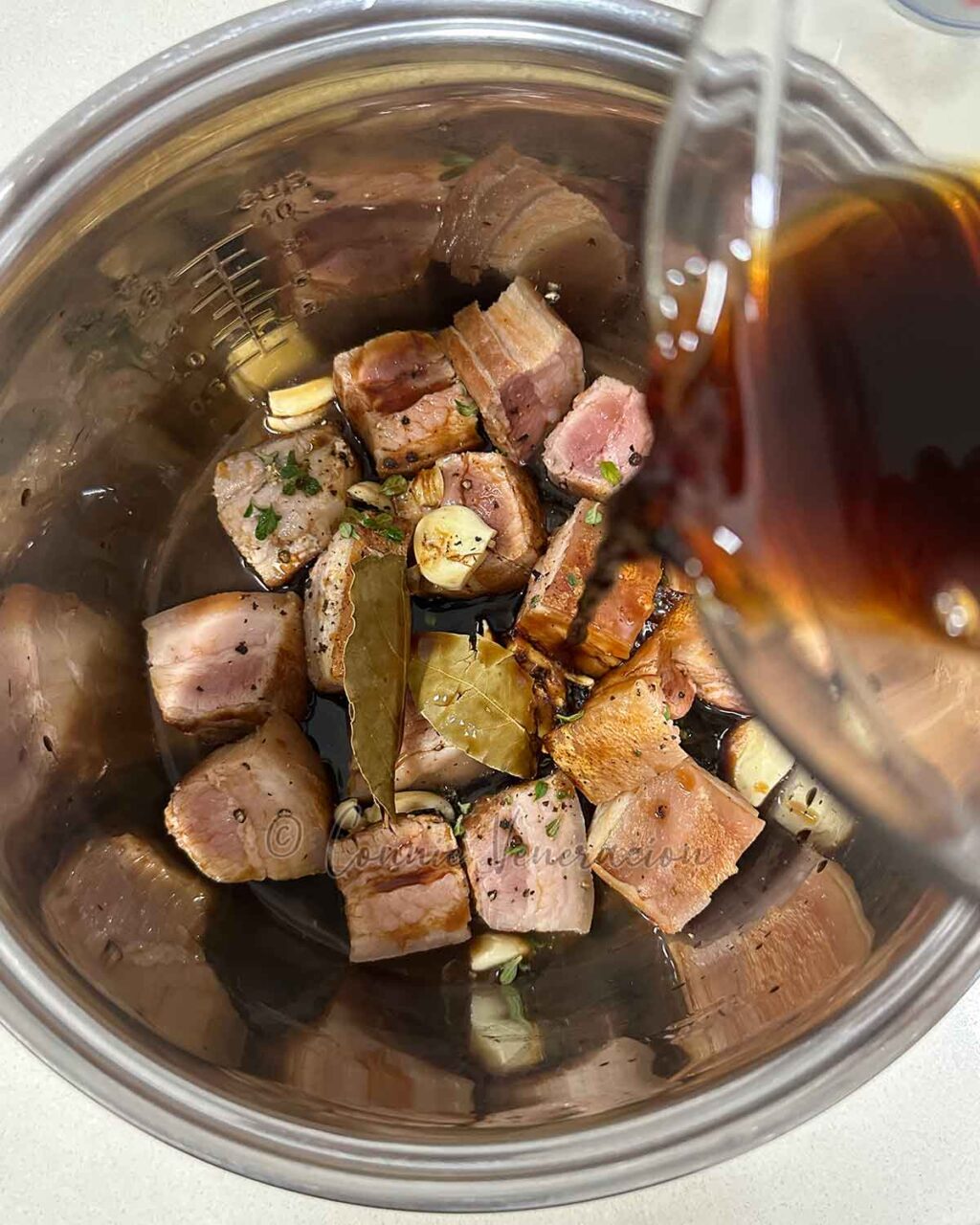 Pouring vinegar and soy sauce over cubes of browned pork belly with oregano, garlic, pepper and bay leaves in the slow cooker