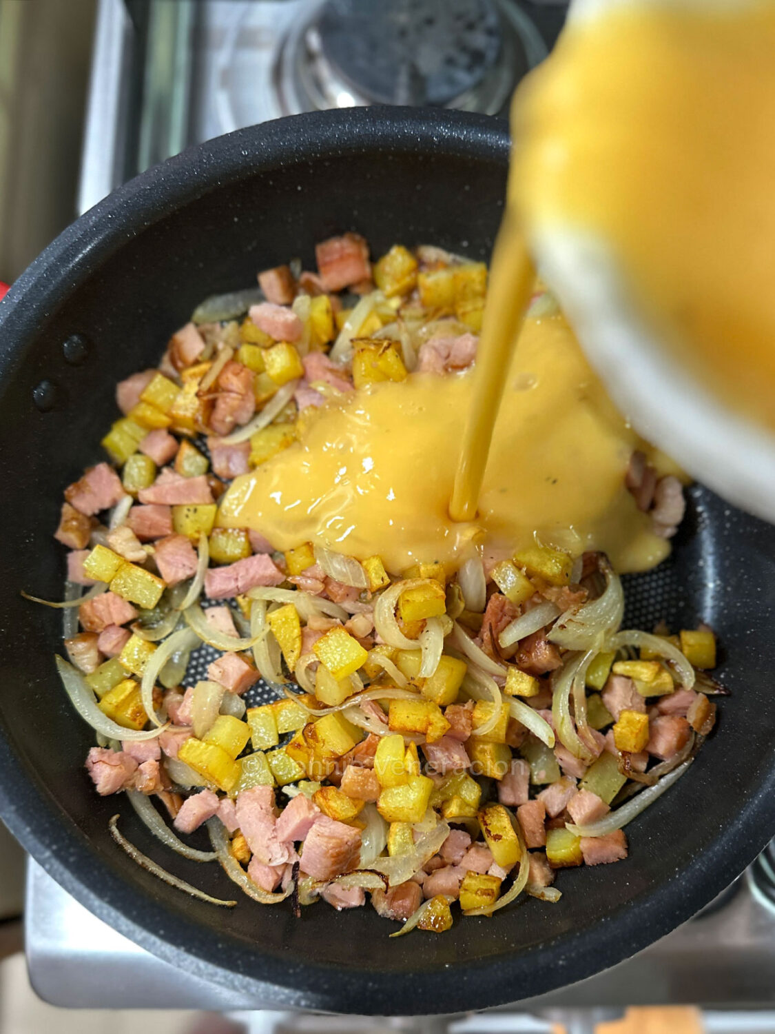 Pouring eggs over potatoes, ham and onions in frying pan
